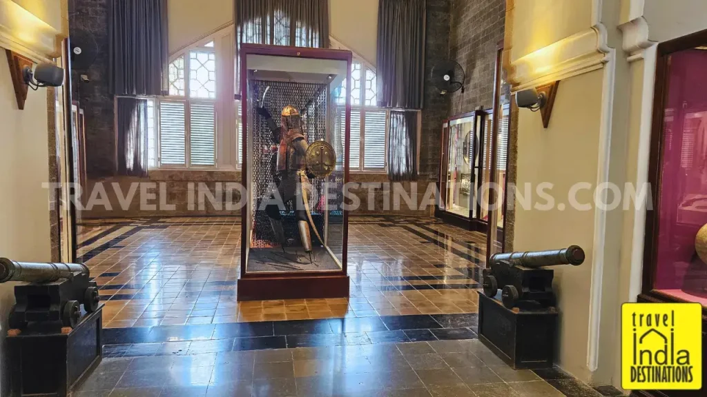 Arms and Armour gallery at Chhatrapati Shivaji Maharaj Vastu Sangrahalaya, featuring a full warrior costume display flanked by cannons, lit by soft afternoon sunlight from large windows.