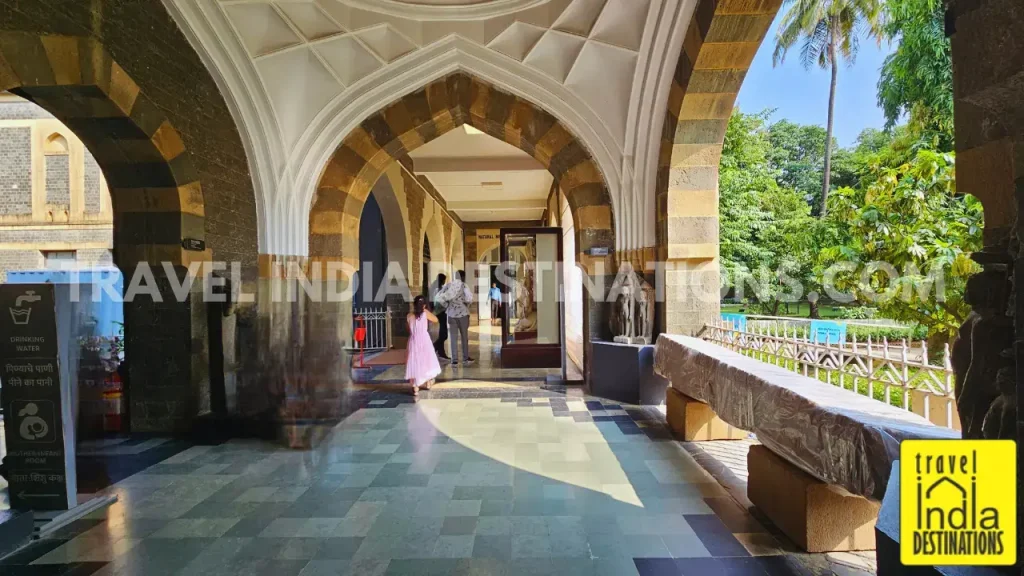 A wide corridor on the ground floor of Chhatrapati Shivaji Maharaj Vastu Sangrahalaya, framed by basalt and Kurla stone arches, leading toward the Natural History section.